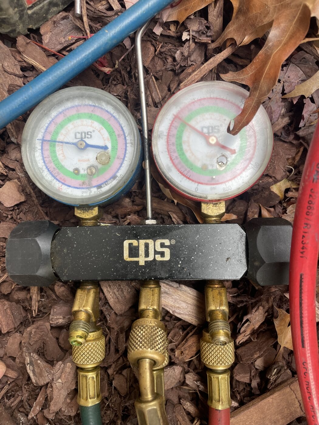 Two CPS pressure gauges connected to hoses, resting on mulch with a dry leaf nearby. The dials show different pressure readings, commonly used by an HVAC contractor for system diagnostics and maintenance.