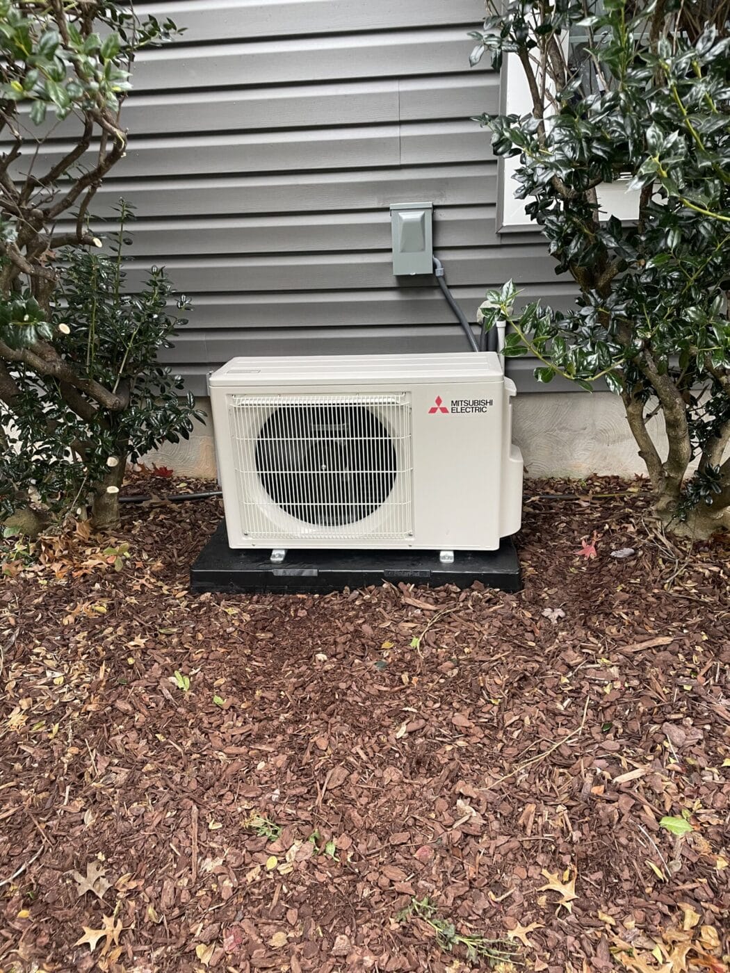 Outdoor air conditioning unit installed next to a house, surrounded by mulch and shrubs.