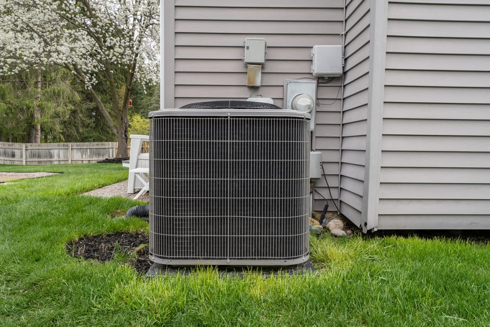 An expertly done AC installation features an outdoor air conditioning unit next to the siding of a house, surrounded by lush grass and a blooming tree in the background.