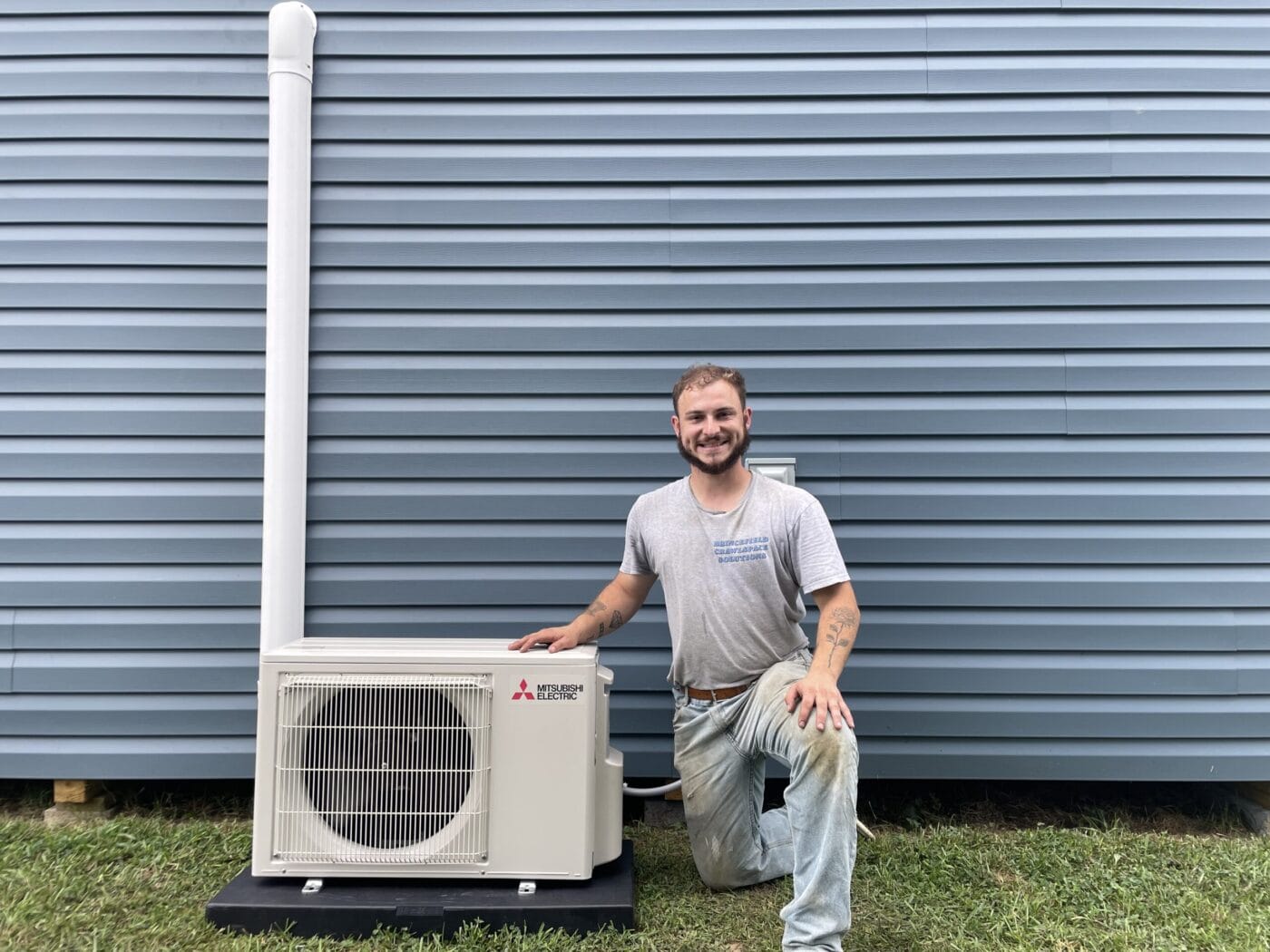 A person kneels beside an installed outdoor air conditioning unit mounted on a platform in front of a blue-paneled wall.