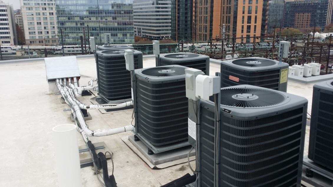 Rooftop with multiple large air conditioning units connected by visible piping, surrounded by urban buildings in the background.