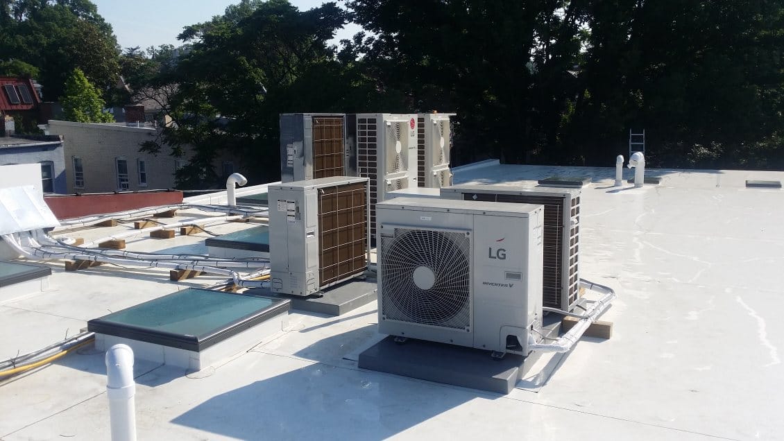 Rooftop with multiple air conditioning units, some labeled LG, and vents under a clear sky with surrounding trees visible.