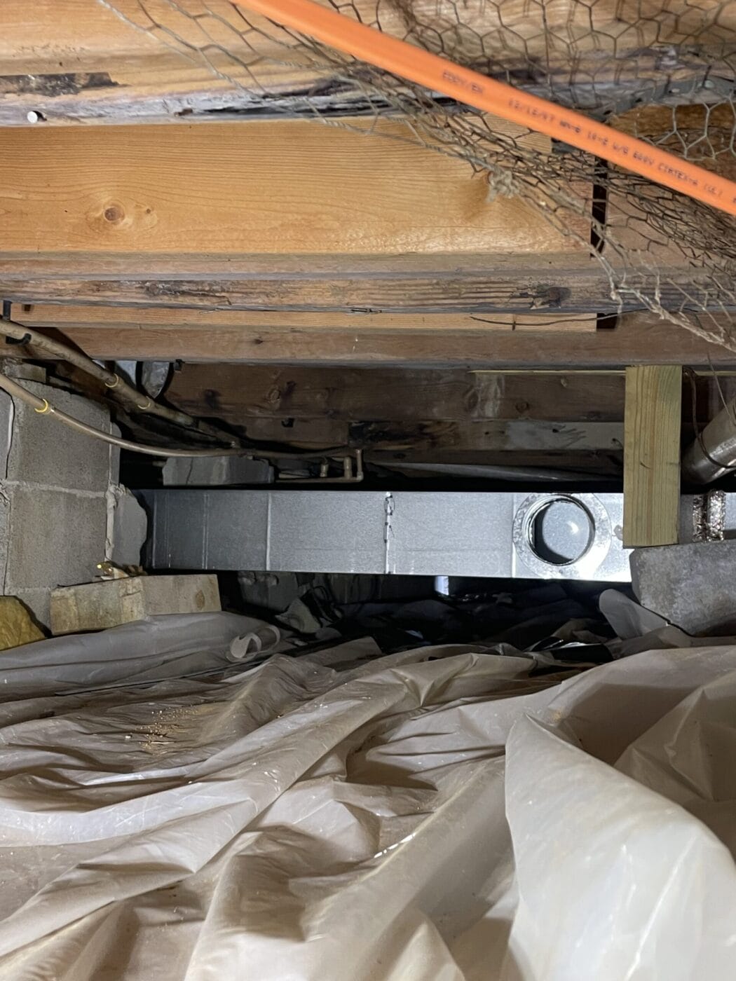 A narrow crawl space under a house with exposed wooden beams, ducts, and plastic sheeting on the ground. An orange cable runs near the ceiling.