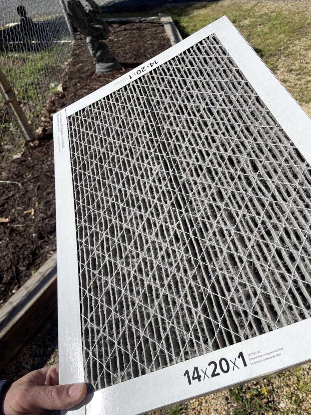 A hand holds a dirty air filter labeled 14x20x1 near a garden area.