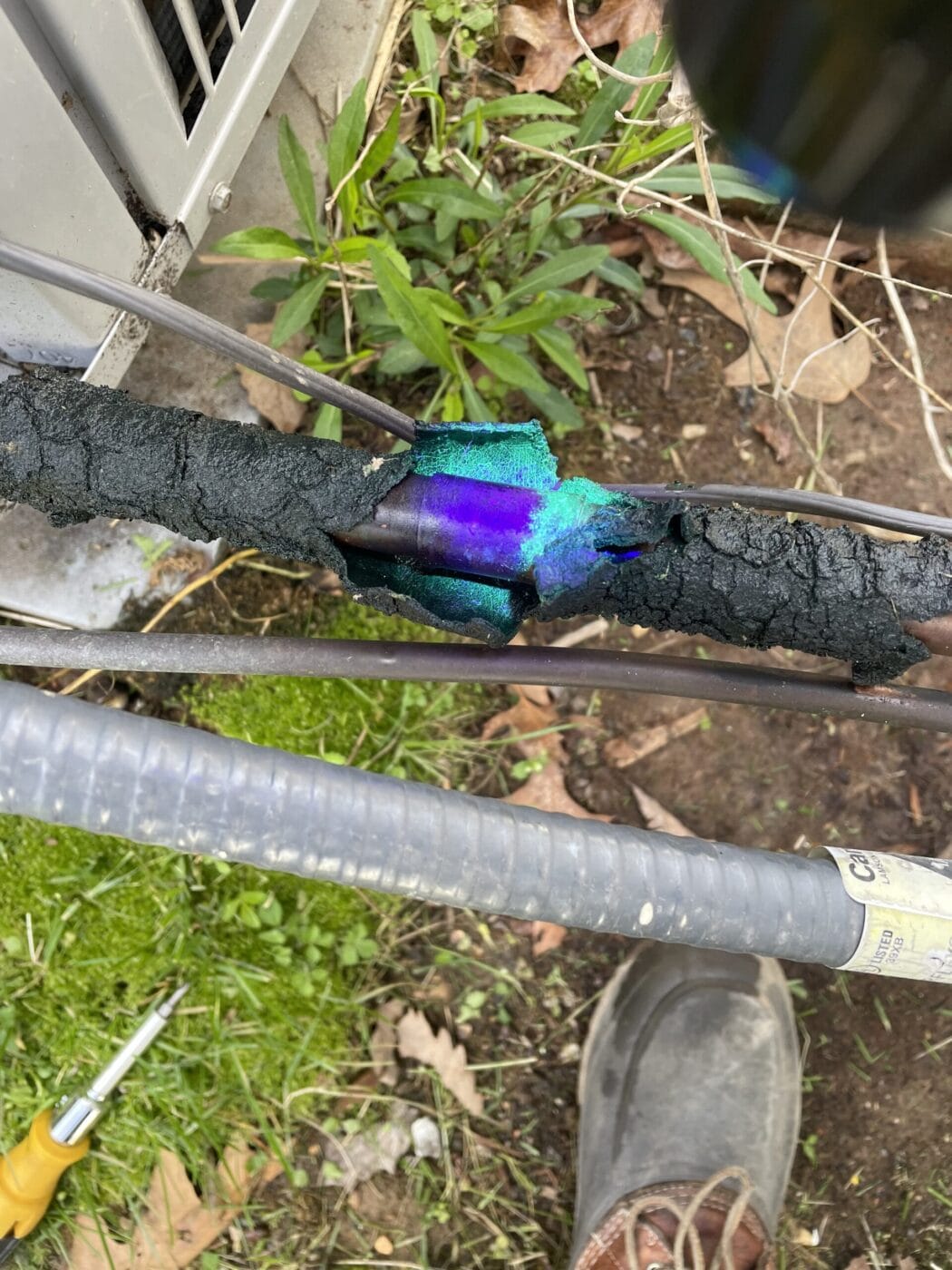 A damaged cable with burnt insulation showing exposed sections, surrounded by grass and foliage. A tool and shoe are partially visible nearby.