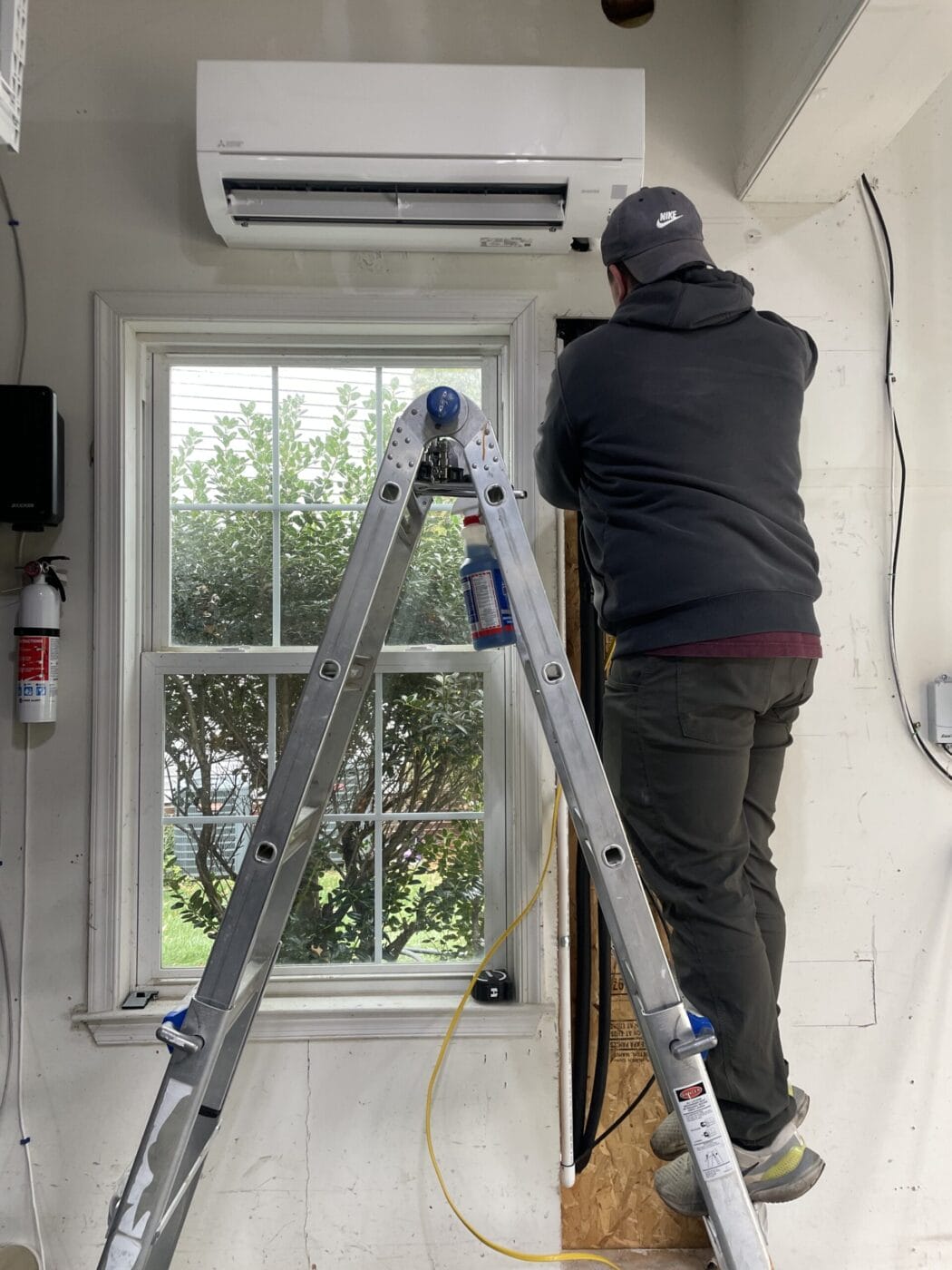 On a ladder, a person expertly installs an air conditioning unit near the window inside the room, seamlessly integrating it as part of an efficient heating installation system.