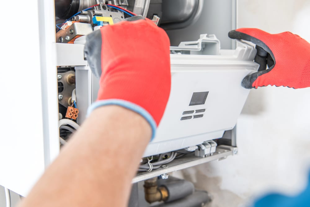 An HVAC contractor wearing red gloves removes the front panel of a gas boiler, exposing internal components for maintenance or repair.
