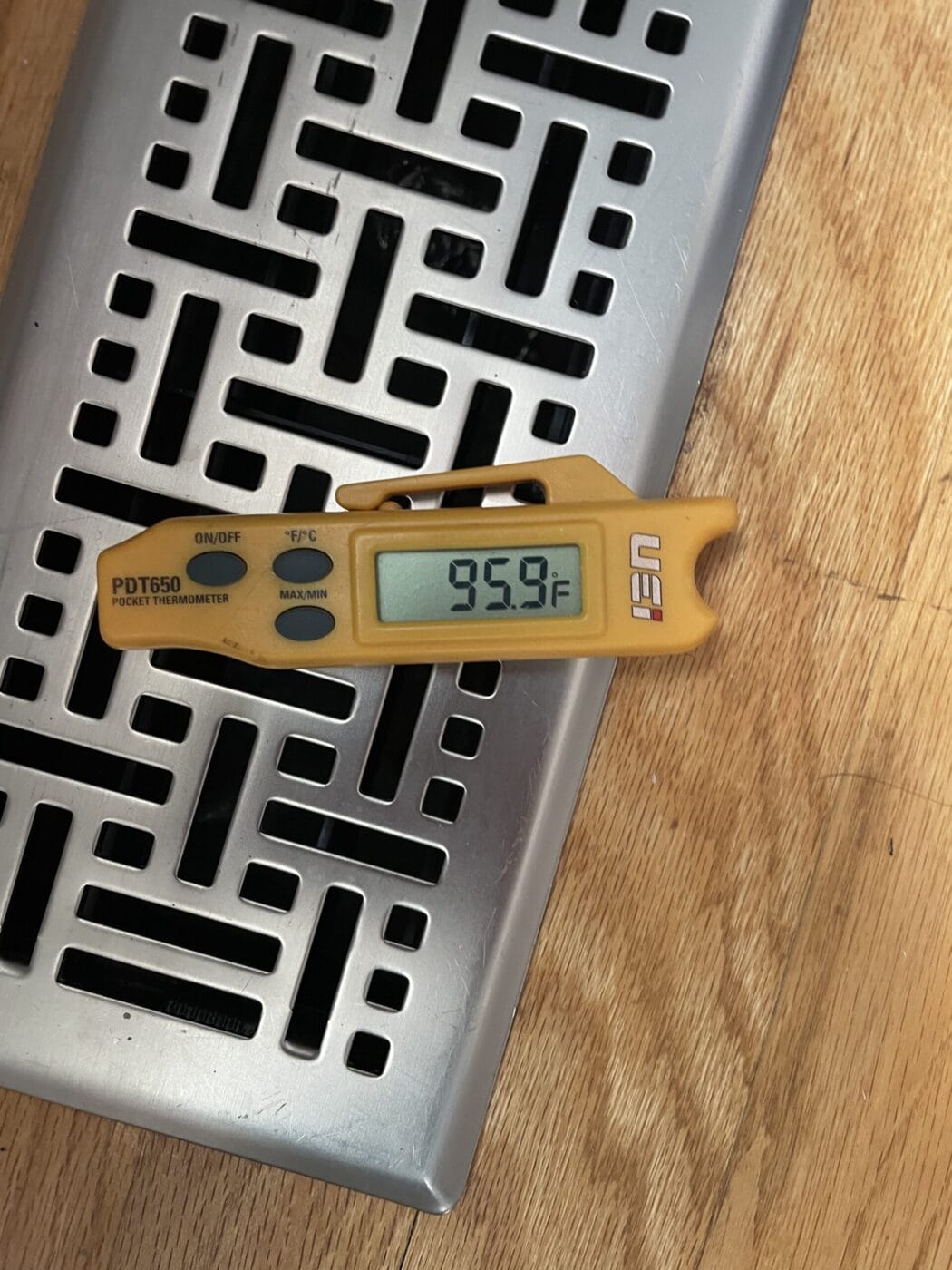 A digital thermometer showing 95.9°F rests on a metal vent cover with a geometric pattern, placed on a wooden surface.