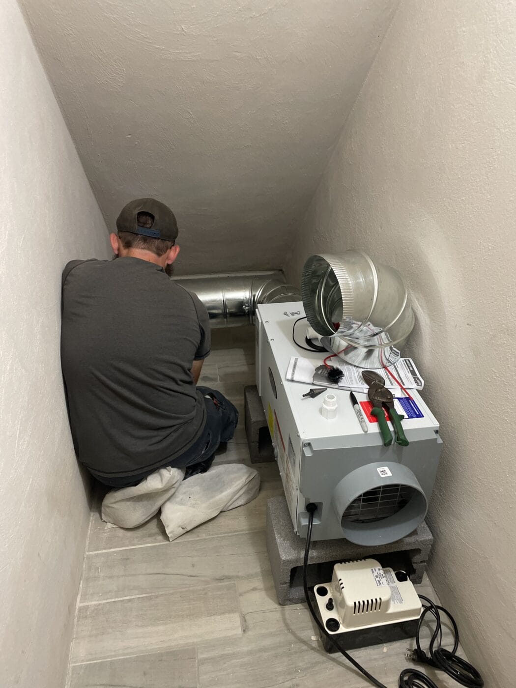 An HVAC contractor in a gray shirt and cap works on installing ductwork connected to a ventilation unit in a small, cramped space, surrounded by tools and equipment.