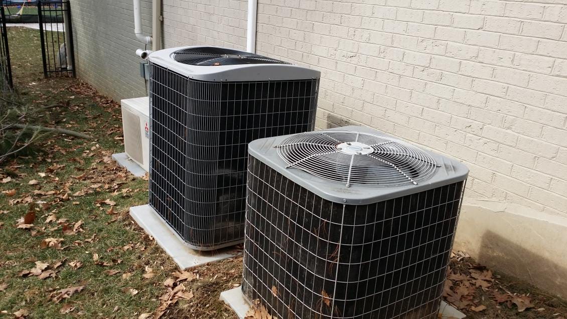 Three outdoor air conditioning units, part of a recent heating installation, are placed beside a brick building, surrounded by fallen leaves on the grass.