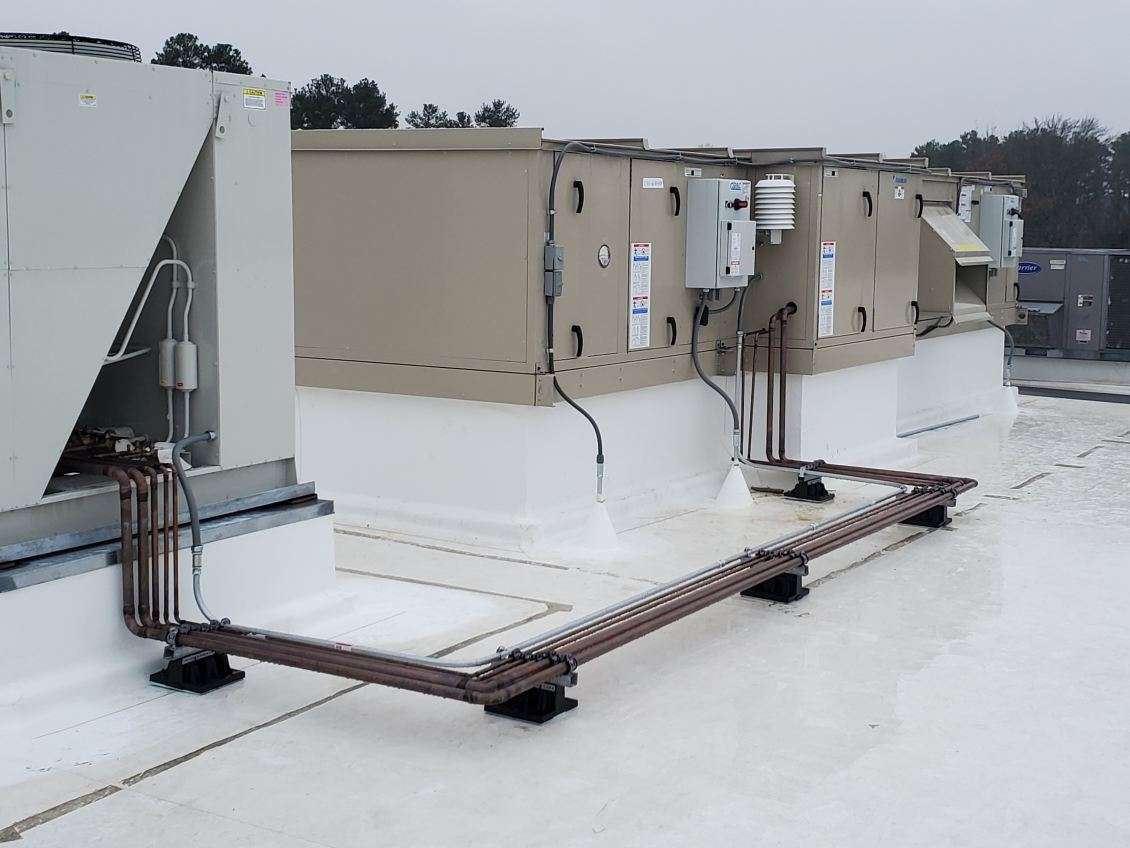 Rooftop view of industrial HVAC units with connected pipes on a flat white surface. Trees are visible in the background under a cloudy sky.